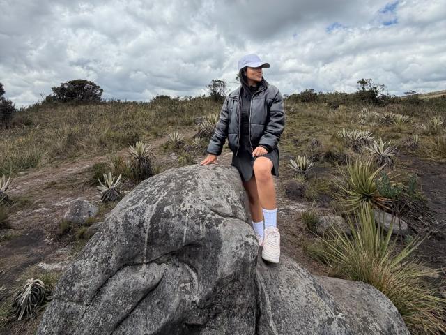 MajoJones, ein Mann mit einer schwarzen Jacke und Mütze, sitzt auf einem großen, grauen Felsen in einer grasbewachsenen, felsigen Landschaft. Er trägt weiße Socken und weiße Turnschuhe, die Beine sind gespreizt. Bewölkter Himmel..