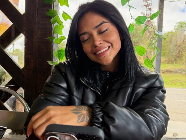 MajoJones mit geschlossenen Augen und einem breiten Lächeln, in einer schwarzen Lederjacke, mit einem Tattoo am rechten Handgelenk, hält ein Smartphone, grün. Blätter im Hintergrund, Sonnenlicht, das hindurchscheint, entspannt und glücklich..