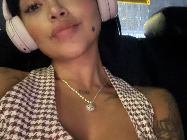 Primer plano de MajoJones, vistiendo una blusa de cuadros blancos y marrones, auriculares blancos, un collar dorado con un dije en forma de corazón. Tatuaje en el hombro derecho. Labios ligeramente entreabiertos, expresión sensual, iluminación tenue, interior de un coche..