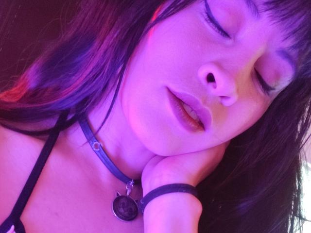 Close-up van IssisDiosa met paars en blauw licht, ogen gesloten, lippen lichtjes open, een zwarte choker met een hanger. Hand rustend op de wang. Haar donkere haar heeft roze highlights. Verdomd sexy..