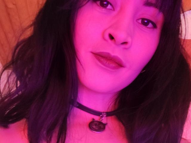 Close-up selfie van IssisDiosa, een Aziatische vrouw met huid met een roze tint, zwart haar en een zwarte choker met een hanger. Haar lippen zijn lichtjes open, haar ogen half gesloten, met een suggestieve uitdrukking..