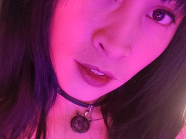 Primo piano di IssisDiosa con illuminazione rosa e viola, capelli neri e una collana nera con un ciondolo circolare. Le sue labbra sono leggermente socchiuse, mostrando i denti. Concentriamoci sul suo viso..