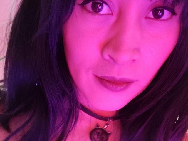 Close-up of IssisDiosa, an Asian woman with long black hair, pink-tinged skin, and dark eyes. She wears a black choker with a pendant, looking directly at the camera with a slightly open mouth. Fucking intense gaze.