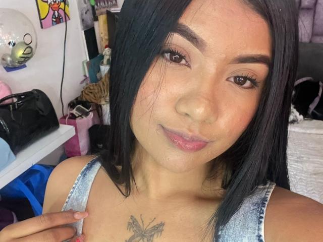 Close-up foto van CamilaDesire met lang zwart haar, lichtbruine huid en een vlindertatoeage op haar borst. Ze draagt een grijze tanktop en kijkt met een lichte glimlach naar de camera. Haar rechterhand raakt haar borst. Helder licht accentueert haar gladde huid. Rommel in de slaapkamer op de achtergrond.