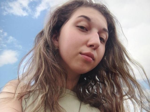 Primer plano de Sanella con el cabello castaño despeinado, piel clara y una expresión ligeramente altanera, mirando hacia abajo a la cámara. Cielo azul con nubes en el fondo, que enfatiza un ambiente casual y ligeramente provocador..