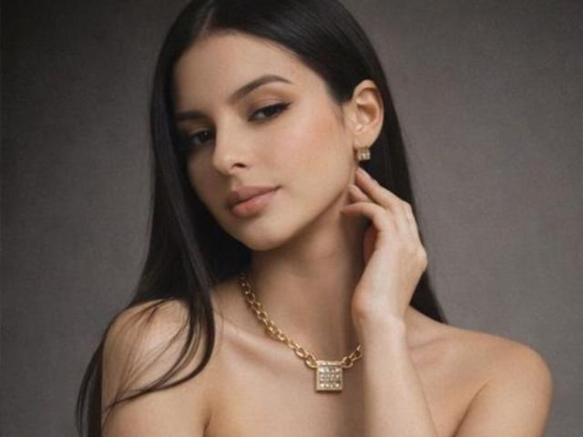 Photograph of IsabellaJames, dark-haired woman with light skin, wearing a gold chain necklace with a square pendant, hand touching neck, sultry expression, soft lighting, grey background, sexy vibe.