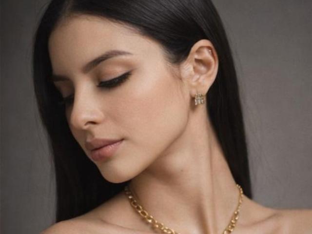 Close-up photo of IsabellaJames, dark-haired woman with closed eyes, wearing gold chain necklace and earrings, head slightly tilted down, soft skin, subtle makeup, grey background, sensual and calm expression.