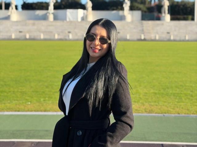IsabellaJames, an attractive woman with long black hair, wearing a black coat over a white top, stands confidently in front of a grassy field. Sunglasses, red lips, and a sexy smile.