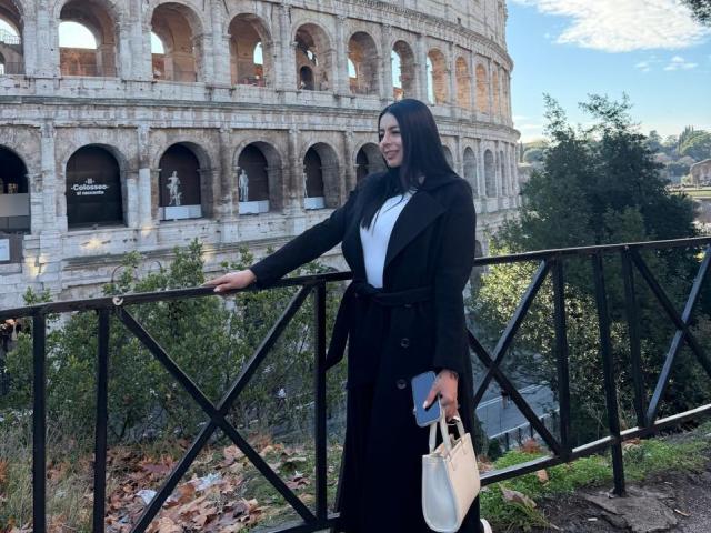 IsabellaJames, with long black hair, stands on a black iron bridge, leaning on the railing. She wears a black blazer over a white top, holding a white handbag. Colosseum in background.