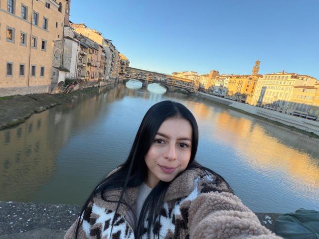 IsabellaJames, with long black hair, stands by a river in a European city, wearing a brown and white patterned coat. Buildings reflect in the water, with a bridge in the background. Fucking beautiful sunset.