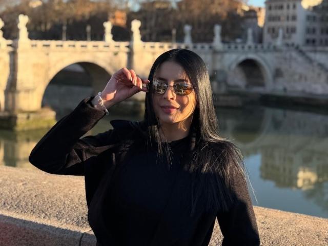 IsabellaJames stands in front of an ornate stone bridge, wearing a black long-sleeve top and reflective sunglasses, adjusting them with her right hand. Her long black hair cascades over her shoulders.