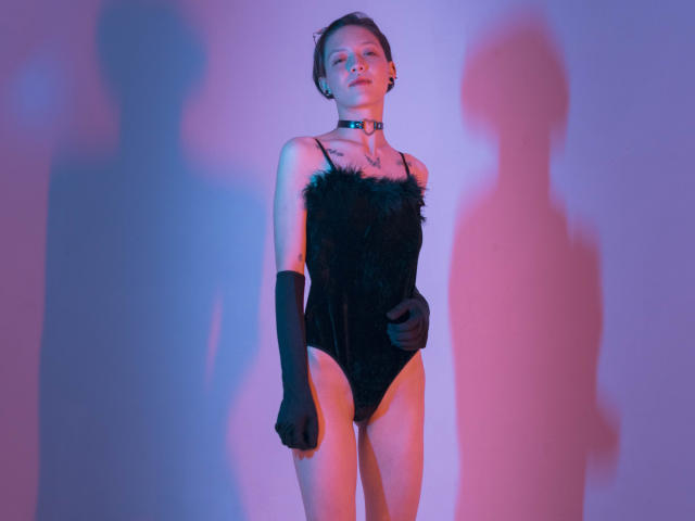 LissyLuxe steht im Mittelpunkt eines Fotos, trägt einen schwarzen, mit Federn besetzten Body und schwarze Handschuhe und wird von blauem Licht beleuchtet. Rosa Beleuchtung.
ihr Schatten fällt nach rechts. Verdammt heiß..
