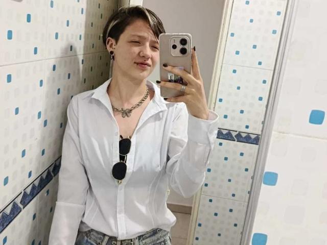 LissyLuxe, con i capelli castani corti, indossa una camicia bianca con bottoni e jeans blu, ed è in piedi in un bagno piastrellato. Selfie allo specchio scattato con un iPhone. Gli occhiali da sole sono appesi alla camicia
.