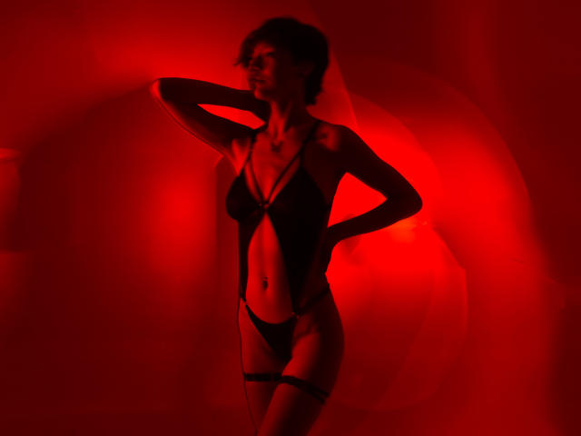 LissyLuxe, silhouet afgebeeld, met kort krullend haar, draagt zwarte, open lingerie, met haar hand achter haar hoofd, en staat zelfverzekerd. Een rode neon achtergrond creëert een intense, erotische sfeer. De schaduwen benadrukken haar gespierde lichaam..
