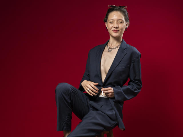 A LissyLuxe está posicionada contra um fundo vermelho sólido, vestindo um terno preto com uma camisa aberta, revelando o peito nu. Ela segura o cinto do traje, com as pernas cruzadas, transmitindo uma aura confiante e sensual..