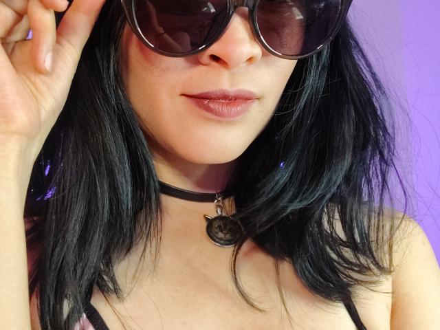 Close-up de IssisDiosa com cabelo preto, usando óculos de sol grandes e escuros, um colar preto com um pingente e uma blusa preta. Ela tem lábios cheios com batom rosa e um leve sorriso. Incrivelmente sexy..