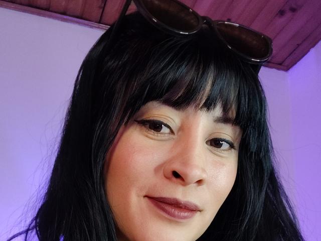 Nahaufnahme eines Fotos von IssisDiosa, einer jungen Frau mit langen, schwarzen Haaren, Pony und braunen Augen. Sie hat ein leichtes Lächeln, trägt eine dunkle Sonnenbrille auf dem Kopf, und über ihr befindet sich eine Holzbalkendecke..