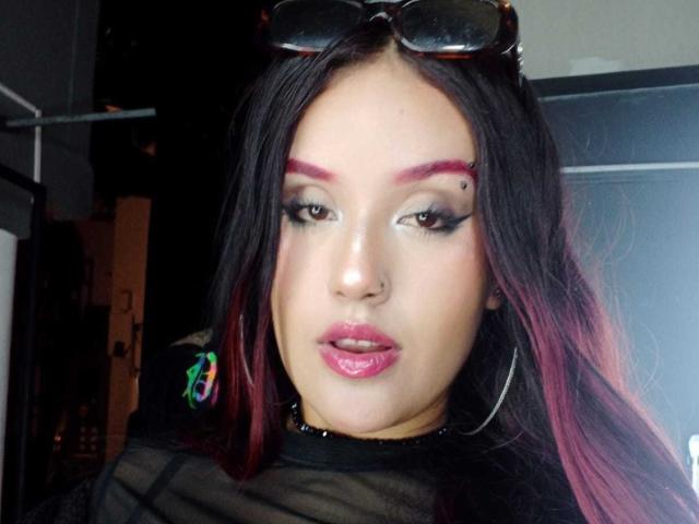 Nahaufnahme-Selfie von MiaDesire, einer kurvigen Frau mit langen, schwarzen und pinken Haaren, starkem Eyeliner und pinkem Lippenstift, die trägt mit schwarzen Sonnenbrillen auf dem Kopf, die direkt in die Kamera blickt und einen verführerischen Ausdruck hat..