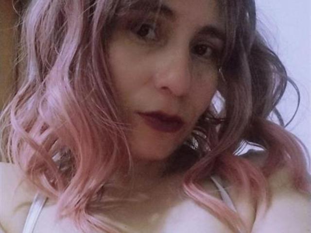 Photo en gros plan de PrettyAlly, avec des cheveux ondulés teintés de rose, du rouge à lèvres foncé et une bouche légèrement entrouverte. Le débardeur blanc de PrettyAlly expose les épaules, créant une ambiance sexy et suggestive..