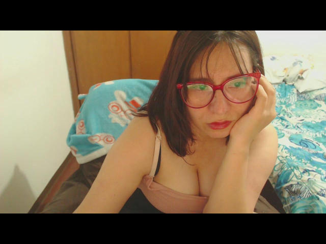 PrettyAlly, a young woman with pale skin and black hair, wears red glasses and a pink tank top, exposing cleavage. She's leaning forward, hand on glasses, looking seductively at the camera.