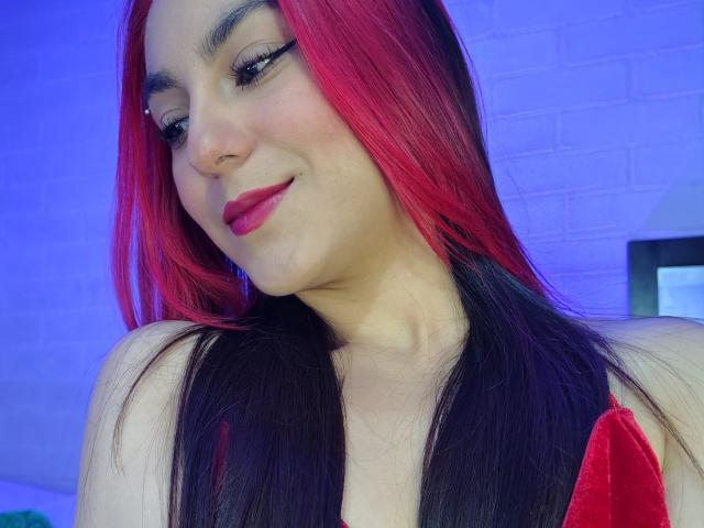 Close-up photo of AmyStreetwise69 with bright red and black hair, wearing red lipstick and a red top, looking down with a slight smile. Blue background. Sexual tension from her relaxed pose.