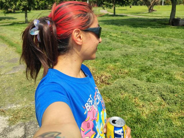 Photograph of AmyStreetwise69, side profile, wearing a blue T-shirt with colorful graphics, red headband, black sunglasses, holding a can, grassy park background, shadowed trees, sunny, casual, relaxed pose.