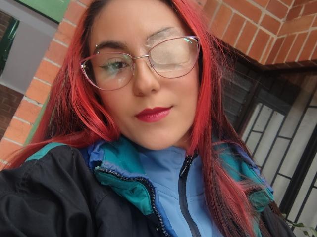 Close-up of AmyStreetwise69 with red hair, wearing clear glasses, blue shirt, and black jacket. She's looking slightly to the side with a subtle smile. Brick wall in background. Sexual tension.