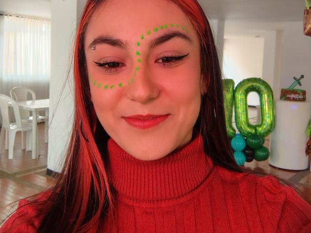 Close-up photo of AmyStreetwise69 with long red hair, wearing a red turtleneck, green face paint dots, and a confident smile. Background includes green balloon letters and white furniture. Fucking cute.