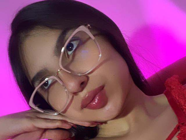 Close-up of MilanMoon, a young woman with dark hair, large transparent glasses, and red lips, sticking out her tongue against a pink and purple neon background. She's wearing a red lace top.