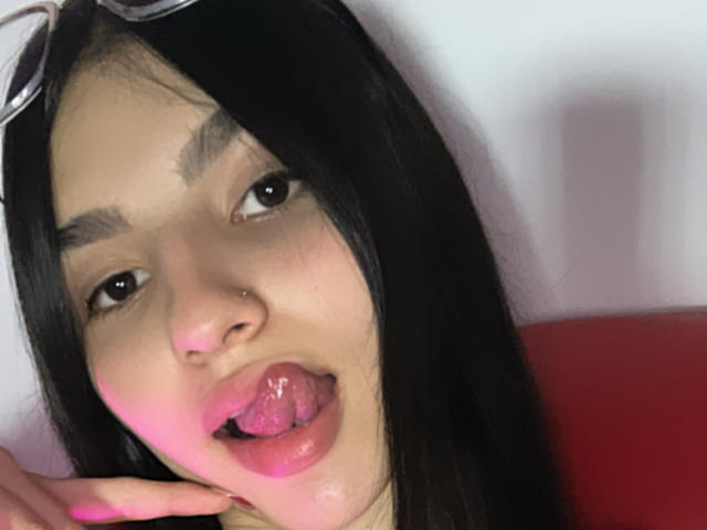 Close-up photo of MilanMoon, a young woman with black hair, thick eyebrows, and a nose piercing. She's sticking out her tongue, with pink gloss and a finger near her mouth. Fucking sexy.