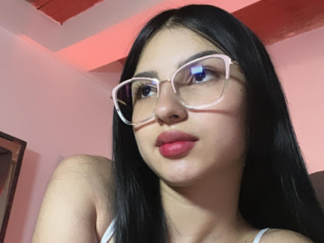 Close-up of MilanMoon, a Latina woman with long black hair, wearing white-framed glasses and red lipstick. She has a neutral expression, slight blush, and pinkish light in the background. Fucking sexy.