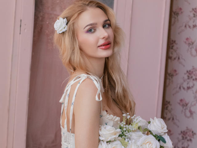 Flora Davis mit blonden Haaren und einem weißen Blumenstrauß im Haar, trägt ein weißes Kleid mit Spitzenbändern und hält einen Blumenstrauß. Weiße Blüten. Sie blickt zurück und lächelt verführerisch..