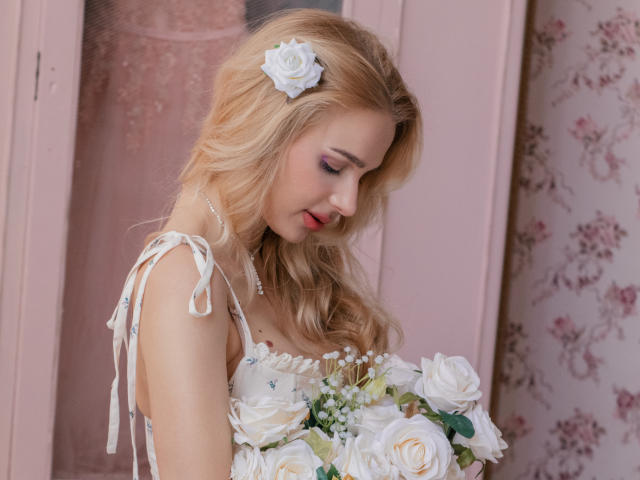 Flora Davis, avec ses longs cheveux blonds ondulés et une fleur blanche dans ses cheveux, portant une robe blanche sans bretelles, tenant une bouquet de roses blanches, yeux fermés, tête inclinée, lumière douce.