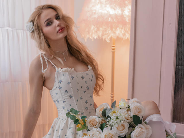 Flora Davis mit langem, gewellten, blondem Haar und einem weißen Kleid mit Blumenmuster, sitzt mit einem Strauß aus weißen und gelben Rosen. Kniestrümpfe, weiches Licht von einer Lampe im Hintergrund. Verdammt heiß..