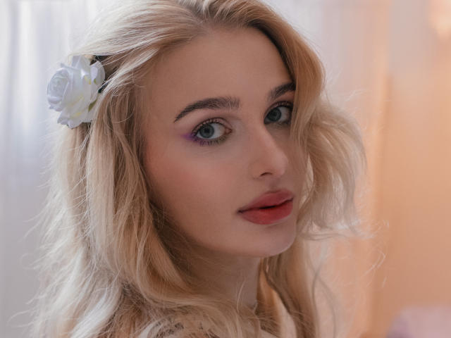 Close-up of FloraDavis, blonde with wavy hair, white flower in hair, blue eyes, purple eyeshadow, red lipstick, soft light background, slightly turned head, sexy gaze, subtle smile, natural makeup.