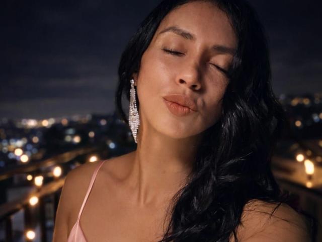 Close-up photograph of BelladonnaLips, eyes closed, lips slightly pursed, long black hair, wearing pink spaghetti straps, large dangling earrings, soft city lights in the background, sensual, night setting.