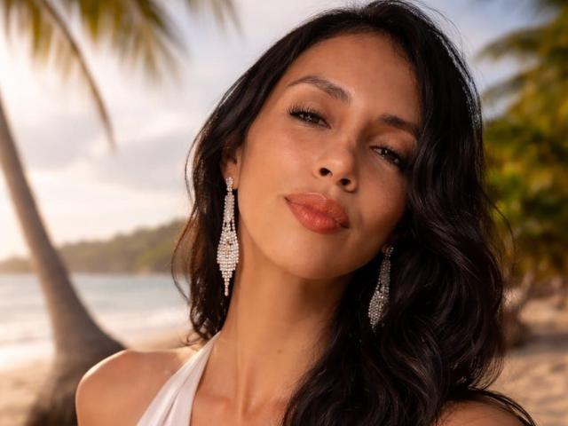 Foto ravvicinata di BelladonnaLips con capelli scuri e ondulati, che indossa un top bianco con spalline e orecchini scintillanti, su una spiaggia illuminata dal sole. con palme sullo sfondo. Sguardo seducente, labbra piene, ambientazione tropicale.