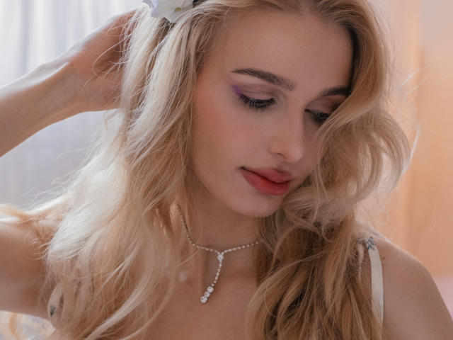 Flora Davis hat langes, welliges blondes Haar, lila Lidschatten und roten Lippenstift. Sie schaut nach unten und berührt mit einer Hand ihr Haar. Sie trägt ein weißes Top mit Trägern und eine Perlenkette. Ihr Ausdruck ist leicht verführerisch..