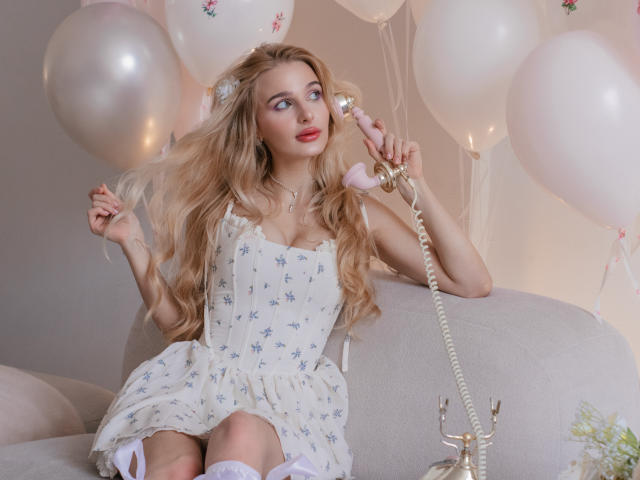 Flora Davis, met lang, golvend blond haar, zit op een grijze bank en houdt een vintage roze telefoon aan haar oor. Ze draagt een witte jurk met bloemen, witte kousen en kijkt verleidelijk. Witte ballonnen en zachte verlichting omringen haar..
