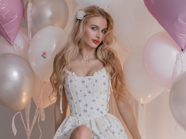 Flora Davis, con lunghi capelli biondi ondulati e rossetto rosso, indossa un abito bianco senza spalline con motivi floreali blu. Circondato da palloncini bianchi e rosa. Ha una clip a forma di fiore tra i capelli..