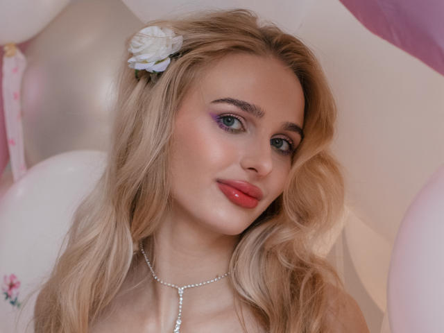 Close-up foto van FloraDavis, een blonde vrouw met golvend haar, die een witte bloem in haar haar draagt en een parelketting, rood. lipsticks, en blauwe oogschaduw. De achtergrond heeft witte ballonnen. Absoluut prachtig..