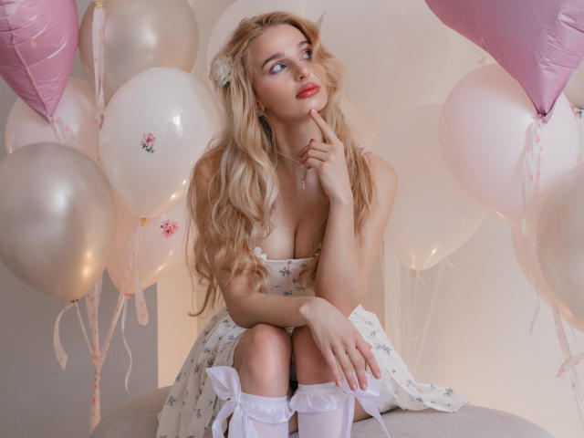 Flora Davis, una donna bionda con lunghi capelli ondulati, è seduta tra palloncini rosa e bianchi, indossando un abito bianco a pois e calze bianche che arrivano al ginocchio. Lei morde il dito in modo seducente, con il rossetto rosso ben visibile 
.