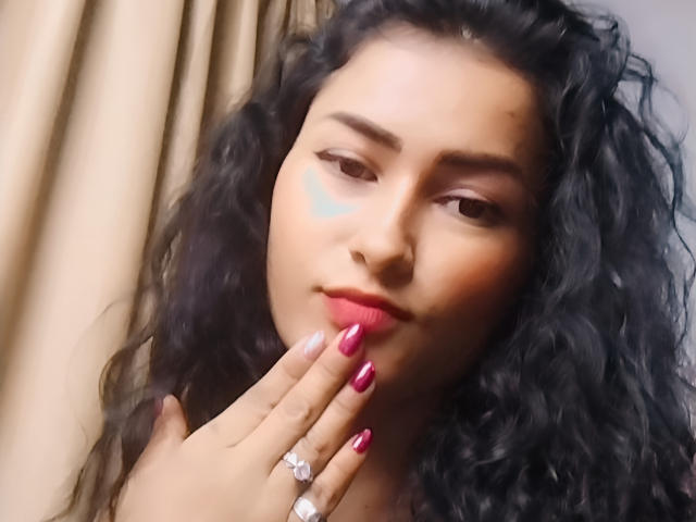 Close-up van DulceLight, een vrouw met krullend zwart haar, rode lippenstift en lange rode nagels, die haar lippen aanraakt met één vinger. Hand, met diamantenringen, die seksuele spanning uitdrukt. Zacht licht.