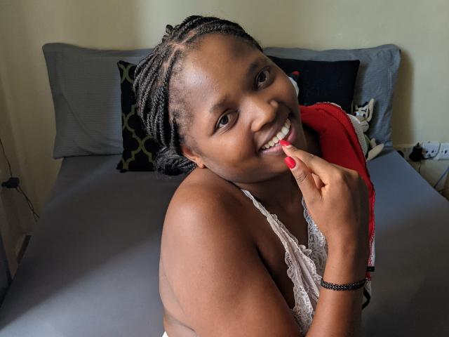 Fotografia de KishaCali, sorrindo de forma sedutora enquanto morde o dedo pintado de vermelho, vestindo uma blusa branca de renda, em um fundo cinza. cama com travesseiros pretos, a luz do sol realçando sua pele escura e tranças..