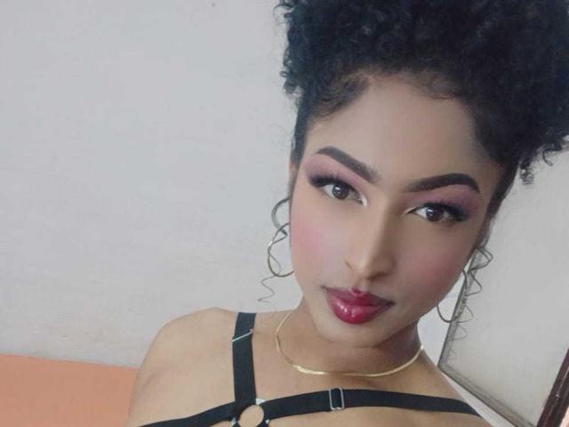 Close-up selfie of BlackMoonTS, an attractive Asian woman with curly black hair, bold eyebrows, pink blush, and red lipstick. She's wearing a black strap harness, looking directly at the camera with a sultry expression.