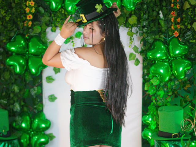 GemmaBellini, long black hair, wearing white off-shoulder top, green velvet skirt, black top hat with shamrock, standing against green leafy backdrop, white wristband, looking back, seductive pose, vibrant colors.