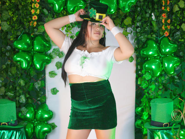 Gemma Bellini está en el centro, con las manos levantadas, ajustándose un sombrero verde. Ella lleva una camiseta corta blanca y una falda verde de terciopelo. El fondo está lleno de globos verdes y plantas. Colores brillantes y vibrantes.