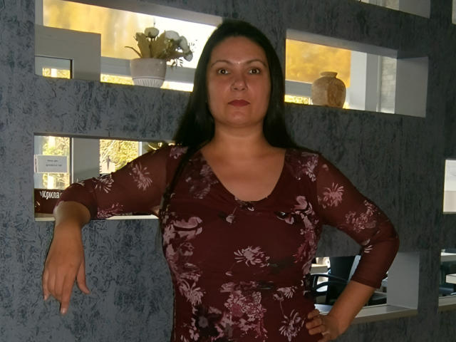 Elenlight has a medium build, brown eyes, and long dark hair. She's wearing a sheer, floral-patterned, maroon dress, with her right arm bent and left hand on her hip.