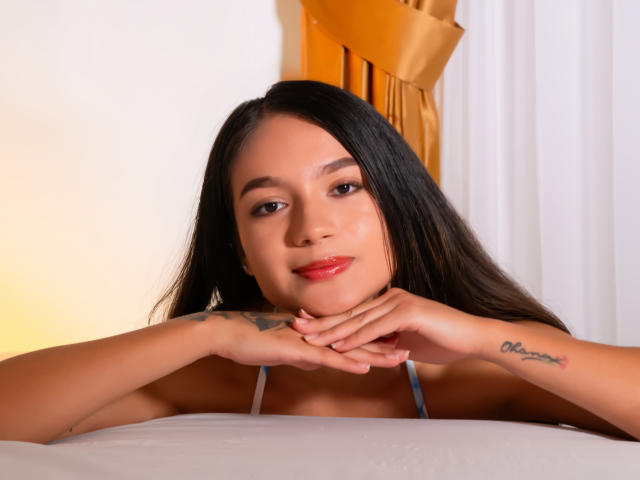 Milly Palmer mit langem, schwarzem Haar, die ihr Kinn in die Hände legt, rotem Lippenstift, einem Tattoo am rechten Handgelenk, einem weißen Bett und einem orangefarbenen Vorhang. Hintergrund, helles Licht, direkter Blick, sexy, entspannt, selbstbewusster Ausdruck..