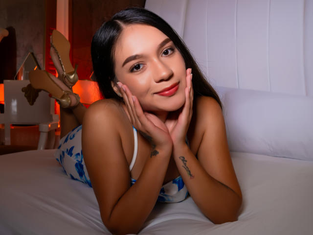 Milly Palmer è sdraiata su un letto, il viso appoggiato sulle mani, con i capelli neri, il rossetto rosso, un abito floreale blu e un tatuaggio sull'avambraccio sinistro. ragazza con tacchi alti, camera da letto con lampada rossa, espressione seducente.